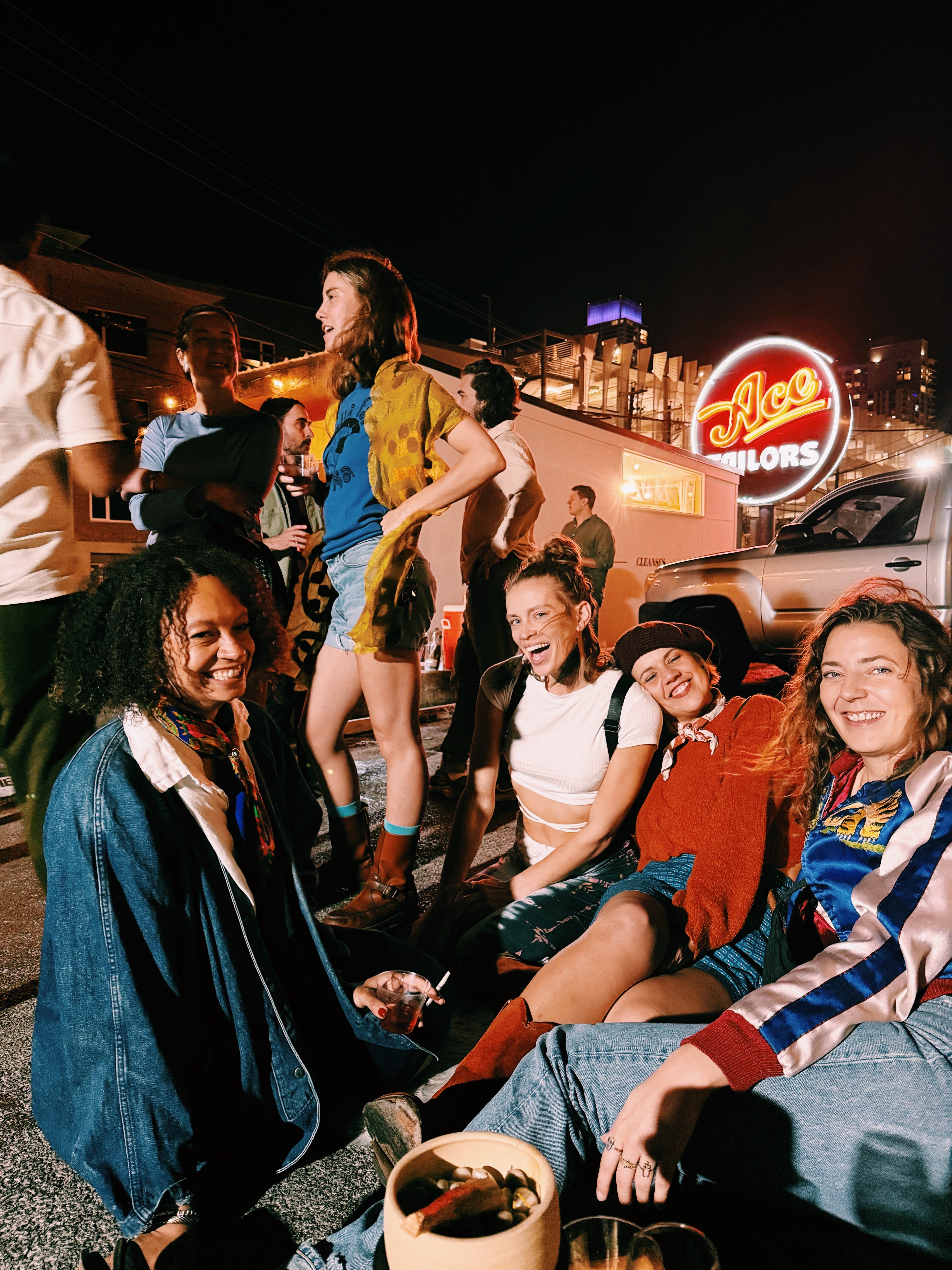 Group of friends sitting together at night in a casual outdoor setting with neon signs in the background. JOCO Easy Hangs event in Austin Texas at Little Tipsy Wine bar. 