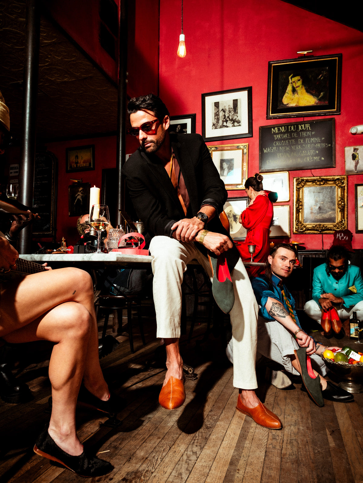 A man in a black blazer, white trousers, and brown JOCO Royal  leather shoes sits on a table in a dimly lit, vintage-style bar with red walls and framed pictures. Other people are visible in the background of the scene.