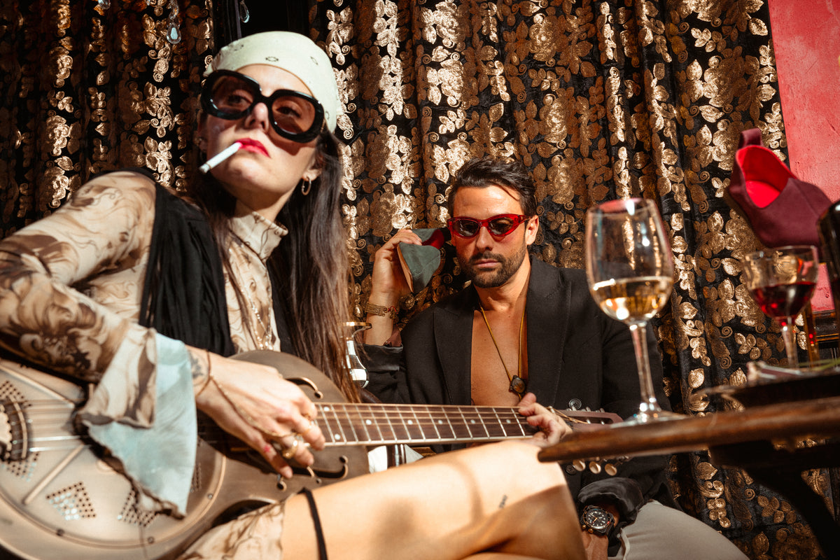 JOCO red suede Mojri loafer on a pedestal beside a woman with a guitar and a man in a gold-lit bar.